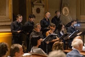 Ensemble vocale in abito nero esegue un brano sacro in chiesa, leggendo gli spartiti su leggii illuminati davanti al pubblico.