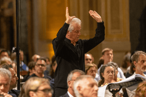 Il direttore, concentrato, indica una sezione del coro durante l’esecuzione, tra il pubblico attento.