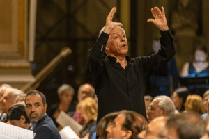 Il direttore apre le braccia per coordinare l’ensemble vocale, mentre il pubblico segue con attenzione all’interno della chiesa.