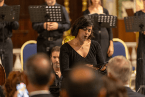 Una cantante solista esegue un brano durante il concerto di musica sacra, circondata dal pubblico attento e dagli altri interpreti sullo sfondo.