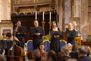 Quattro cantanti, due uomini e due donne, eseguono un brano di musica sacra accompagnati da un ensemble strumentale, sullo sfondo l’altare riccamente decorato.