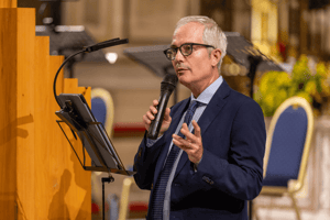 Un uomo in abito blu parla al microfono durante il Concerto di Musica Sacra, rivolgendosi al pubblico all’interno di una chiesa illuminata, con partiture appoggiate su un leggio accanto a lui.
