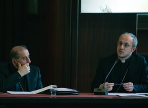 Mons. Mario Delpini saluta con una stretta di mano un sacerdote durante la conferenza; sullo sfondo un uomo in abito elegante prende parte alla conversazione.