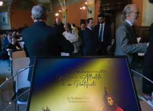 Monitor con il titolo dell’evento in primo piano; in secondo piano, il pubblico si prepara alla conferenza nella sala affrescata.