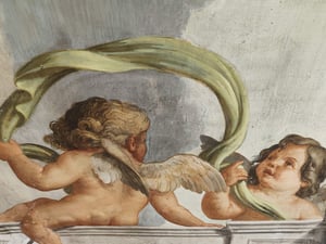 Dettaglio di affresco raffigurante due putti alati: uno di spalle e uno frontale, che sollevano e intrecciano un drappo verde su sfondo celeste