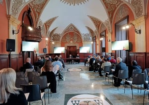 Sala conferenze storica con soffitti affrescati e pareti decorate, dove un gruppo di persone è seduto ad assistere a un incontro o convegno; due relatori sono seduti a un tavolo sul palco, con schermi laterali che proiettano immagini