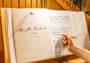 Primo piano di uno spartito aperto sul leggio di un organo, con il titolo ‘Messa Papa Benedetto XIII a 16 voci’ di Giuseppe Ottavio Pitoni; una mano tiene una matita puntata sulle note musicali